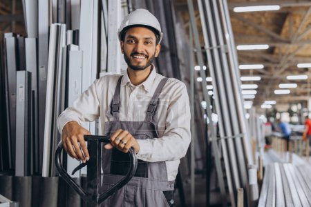 Téléchargez les photos : Portrait d'un heureux travailleur hindou portant un casque blanc et une salopette tenant un camion hydraulique sur fond d'usine et de cadres en aluminium. - en image libre de droit