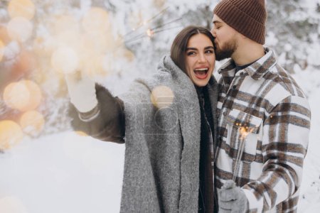 Téléchargez les photos : Couple romantique souriant en bonnets tricotés posant avec des étincelles à la forêt d'hiver, tenant les lumières du Bengale dans les mains, célébrant ensemble les vacances de Noël, Copier l'espace - en image libre de droit