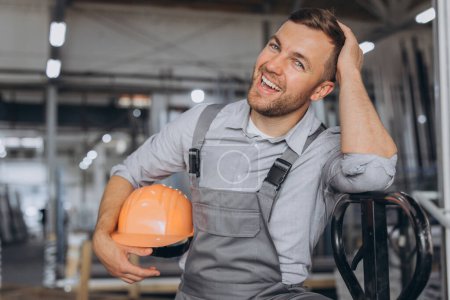 Téléchargez les photos : Portrait d'un travailleur heureux portant un casque orange et une salopette tenant un camion hydraulique sur fond d'usine et de cadres en aluminium. - en image libre de droit