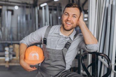 Photo for Portrait of a happy worker in a orange hard hat and overalls holding a hydraulic truck against a background of a factory and aluminum frames. - Royalty Free Image