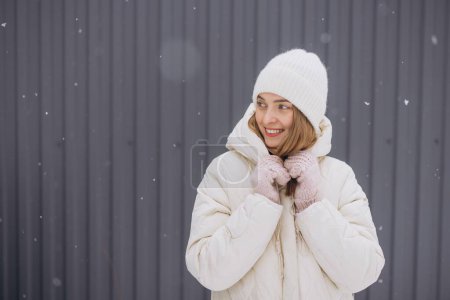 Téléchargez les photos : Une femme heureuse en mitaines tricotées pose sur un fond de mur gris en hiver dans une chute de neige - en image libre de droit