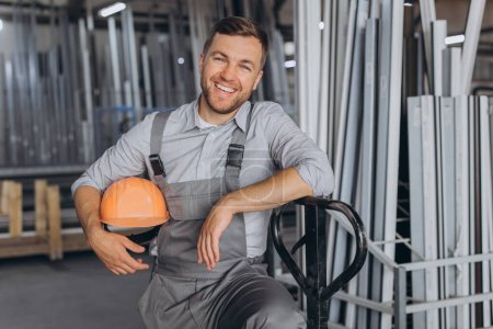 Téléchargez les photos : Portrait d'un travailleur heureux portant un casque orange et une salopette tenant un camion hydraulique sur fond d'usine et de cadres en aluminium. - en image libre de droit