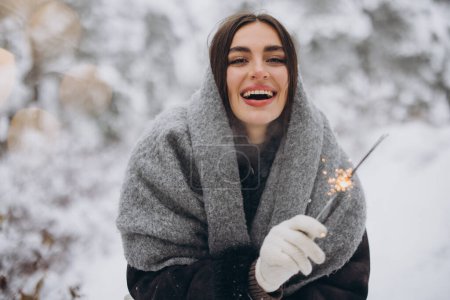 Téléchargez les photos : Portrait de jolie femme heureuse en écharpe tricotée et mitaines tenant scintillant et posant dans la forêt enneigée - en image libre de droit