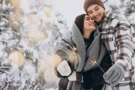Foto de Pareja romántica sonriente en sombreros de punto posando con chispas en el bosque de invierno, sosteniendo luces de bengala en las manos, celebrando vacaciones de Navidad juntos, Copiar el espacio - Imagen libre de derechos