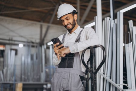 Photo for Portrait of a happy Hindu worker in a white helmet and overalls holding a hydraulic truck and talking on the phone against a background of a factory and aluminum frames. - Royalty Free Image