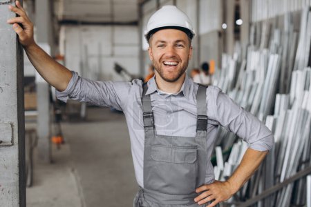 Photo for Portrait of a happy worker in a gray uniform and a white hard hat posing on the background of factory production - Royalty Free Image