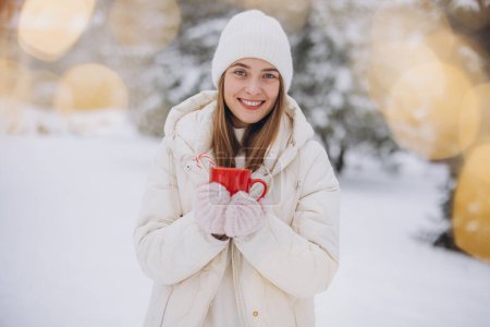 Photo for Cute happy woman in knitted mittens holding hot coffee with candy canes in winter park - Royalty Free Image