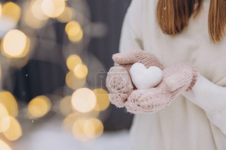 Téléchargez les photos : Les mains féminines en moufles tricotées tiennent un cœur de neige sur un fond d'hiver gris avec des étincelles - en image libre de droit