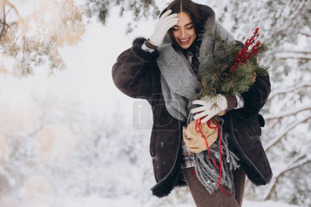 Téléchargez les photos : Portrait de jolie femme heureuse en écharpe tricotée et mitaines tenant bouquet de pin d'hiver et posant dans la forêt enneigée - en image libre de droit