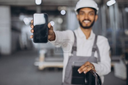 Photo for Portrait of happy hindu worker in white hard hat and overalls holding smartphone and showing blank screen for copy on factory background. - Royalty Free Image