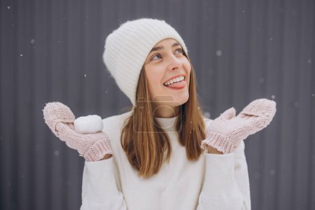 Téléchargez les photos : Femme heureuse en mitaines tricotées essayant d'attraper des flocons de neige avec sa langue sur un fond gris en hiver - en image libre de droit