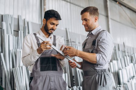 Foto de Dos trabajadores masculinos internacionales en uniforme discuten el proceso de trabajo en una fábrica de ventanas de metal y plástico - Imagen libre de derechos