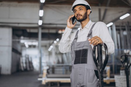 Photo for Portrait of a happy Hindu worker in a white helmet and overalls holding a hydraulic truck and talking on the phone against a background of a factory and aluminum frames. - Royalty Free Image