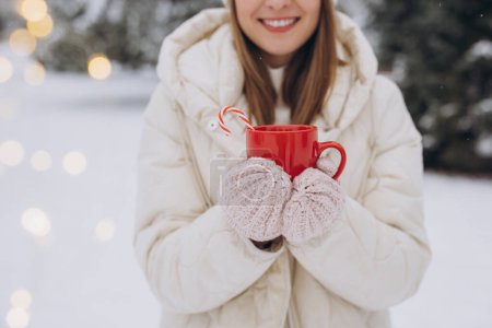 Téléchargez les photos : Mignonne femme heureuse en mitaines tricotées tenant du café chaud avec des cannes à bonbons dans le parc d'hiver - en image libre de droit