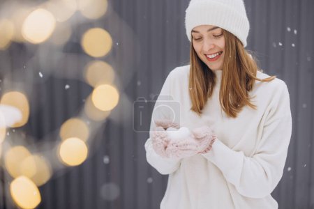 Foto de Mujer feliz en manoplas de punto sosteniendo un corazón de nieve en un día nevado de invierno - Imagen libre de derechos
