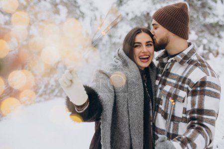 Photo for Smiling Romantic Couple In Knitted Hats Posing With Sparklers At Winter Forest, Holding Bengal Lights In Hands, Celebrating Christmas Holidays Together, Copy Space - Royalty Free Image