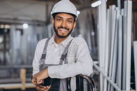 Téléchargez les photos : Portrait d'un heureux travailleur hindou portant un casque blanc et une salopette tenant un camion hydraulique sur fond d'usine et de cadres en aluminium. - en image libre de droit