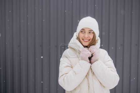 Foto de Una mujer feliz en manoplas de punto posa sobre un fondo de una pared gris en invierno en una nevada - Imagen libre de derechos