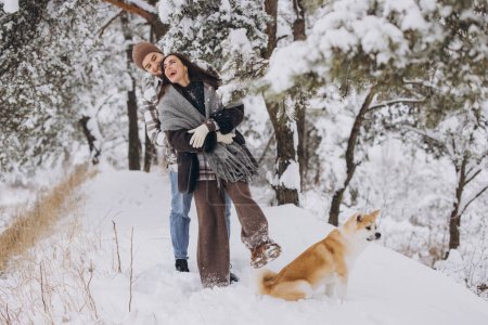 Téléchargez les photos : Joyeux jeune couple marchant avec un chien akita en forêt en hiver et jour neigeux - en image libre de droit