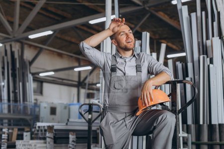 Téléchargez les photos : Portrait de travailleur heureux en chapeau dur orange et salopettes tenant camion hydraulique et allant sur la pause déjeuner essuyant la sueur du front sur fond d'usine. - en image libre de droit