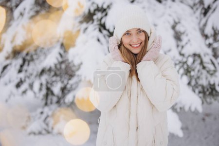 Téléchargez les photos : Une femme heureuse en mitaines tricotées pose sur le fond d'un pin neigeux en hiver - en image libre de droit