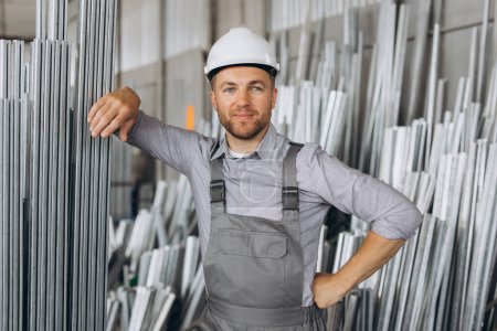 Photo for Happy bearded male factory worker in special uniform and white hard hat holding aluminum frame at production of metal plastic windows and doors - Royalty Free Image