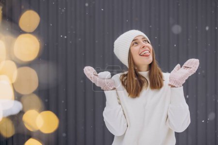 Photo for Happy woman in knitted mittens trying to catch snowflakes with her tongue on a gray background in winter - Royalty Free Image