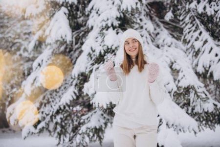 Foto de Mujer feliz en ropa blanca tejida sosteniendo copos de nieve en el fondo de un pino nevado en invierno - Imagen libre de derechos
