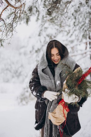 Téléchargez les photos : Portrait de jolie femme heureuse en écharpe tricotée et mitaines tenant bouquet de pin d'hiver et posant dans la forêt enneigée - en image libre de droit