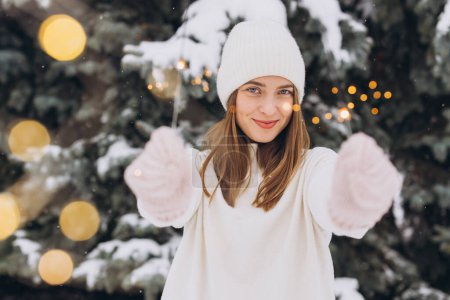 Téléchargez les photos : Portrait de fille souriante portant un chapeau en tricot blanc, une écharpe, des mitaines et un pull avec un éclat en hiver à l'extérieur. Vacances de Noël - en image libre de droit
