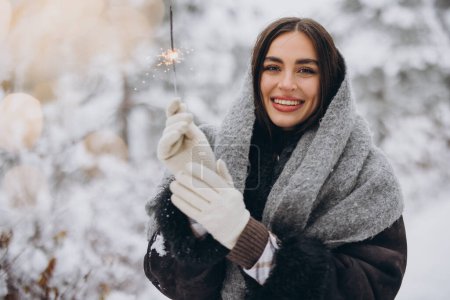 Foto de Retrato de mujer bonita feliz en bufanda de punto y manoplas que sostienen chispeante y posan en el bosque nevado - Imagen libre de derechos