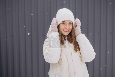 Photo for A happy woman in knitted mittens poses against a background of a gray wall in winter in a snowfall - Royalty Free Image