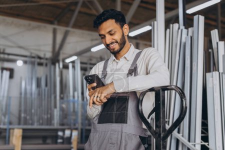 Téléchargez les photos : Portrait d'un travailleur hindou heureux dans un casque blanc et salopettes tenant un camion hydraulique et parlant au téléphone sur un fond d'usine et cadres en aluminium. - en image libre de droit