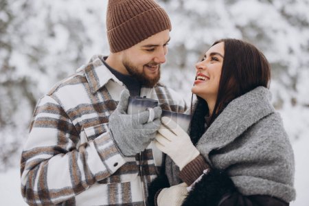 Téléchargez les photos : Heureux beau couple se reposant et buvant du thé chaud dans la forêt en hiver dans les montagnes - en image libre de droit