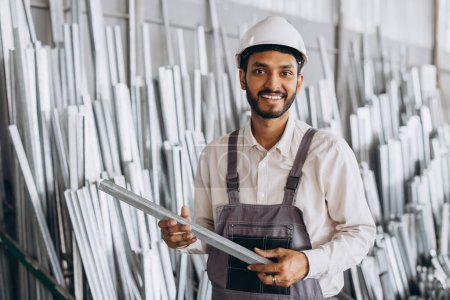 Foto de Trabajador de fábrica masculino indio barbudo feliz en uniforme especial y sombrero duro blanco que sostiene el marco de aluminio en la producción de ventanas y puertas plásticas del metal - Imagen libre de derechos