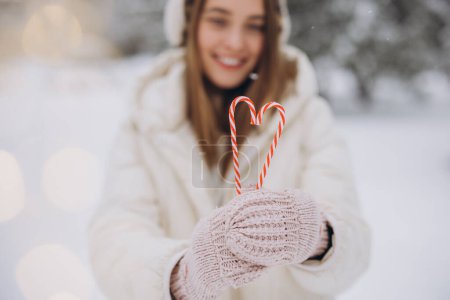 Photo for A cute girl in knitted mittens holds candy canes in the form of a heart in winter park - Royalty Free Image