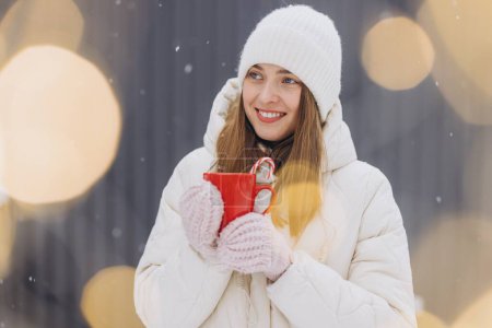 Téléchargez les photos : Mignonne femme heureuse en mitaines tricotées tenant du café chaud avec des cannes à bonbons sur le mur gris - en image libre de droit