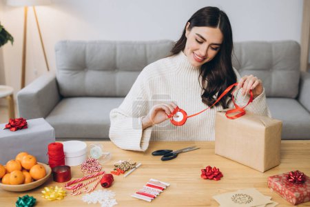 Téléchargez les photos : Joyeux femme en pull tricoté enveloppant cadeaux de Noël en utilisant du papier, des ciseaux et des rubans colorés. Préparatifs de vacances festives. - en image libre de droit