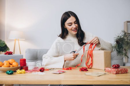 Foto de Mujer feliz en jersey de punto envolviendo regalos de Navidad con papel, tijeras y cintas de colores. Preparativos festivos de vacaciones. - Imagen libre de derechos