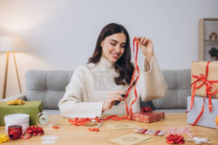 Téléchargez les photos : Joyeux femme en pull tricoté enveloppant cadeaux de Noël en utilisant du papier, des ciseaux et des rubans colorés. Préparatifs de vacances festives. - en image libre de droit