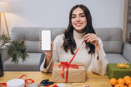 Téléchargez les photos : Belle femme heureuse faisant Nouvel An ou emballage cadeau de Noël tout en tenant fond de téléphone intelligent avec écran blanc avec espace de copie ou se moquer à la maison - en image libre de droit