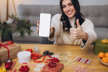 Téléchargez les photos : Belle femme heureuse faisant Nouvel An ou emballage cadeau de Noël tout en tenant fond de téléphone intelligent avec écran blanc avec espace de copie ou se moquer à la maison - en image libre de droit