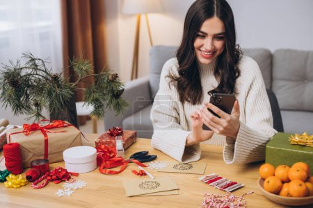 Foto de Hermosa mujer feliz envolviendo regalos de Navidad y el uso de teléfonos inteligentes cerca de la decoración borrosa en casa - Imagen libre de derechos