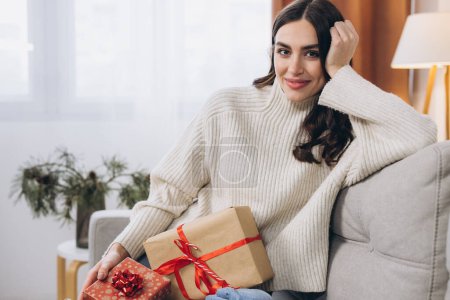 Photo for Beautiful happy woman unwrapping New Year or Christmas presents at home on sofa - Royalty Free Image