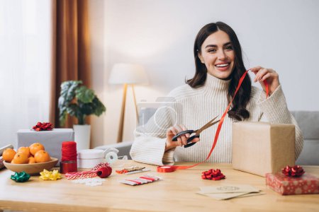 Photo for Happy woman in knitted sweater wrapping Christmas presents using paper, scissors and colorful ribbons. Festive holiday preparations. - Royalty Free Image