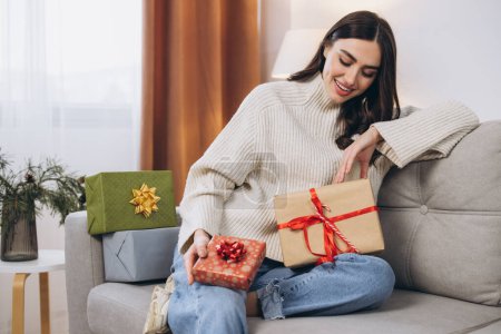 Téléchargez les photos : Belle femme heureuse déballer des cadeaux de Nouvel An ou de Noël à la maison sur le canapé - en image libre de droit