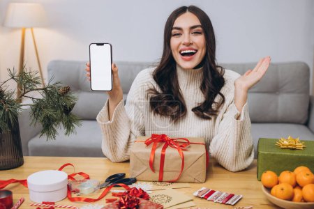 Foto de Hermosa mujer feliz haciendo envoltura de regalo de Año Nuevo o Navidad mientras sostiene el fondo del teléfono inteligente con pantalla en blanco con espacio para copiar o burlarse en casa - Imagen libre de derechos