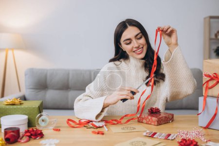 Foto de Mujer feliz en jersey de punto envolviendo regalos de Navidad con papel, tijeras y cintas de colores. Preparativos festivos de vacaciones. - Imagen libre de derechos