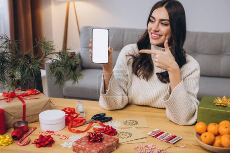 Photo for Beautiful happy woman doing New Year or Christmas gift wrapping while holding smart phone background with blank screen with copy space or mock up at home - Royalty Free Image