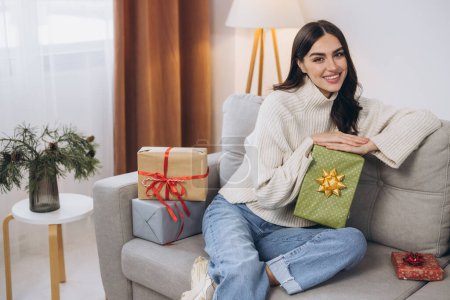 Téléchargez les photos : Belle femme heureuse déballer des cadeaux de Nouvel An ou de Noël à la maison sur le canapé - en image libre de droit